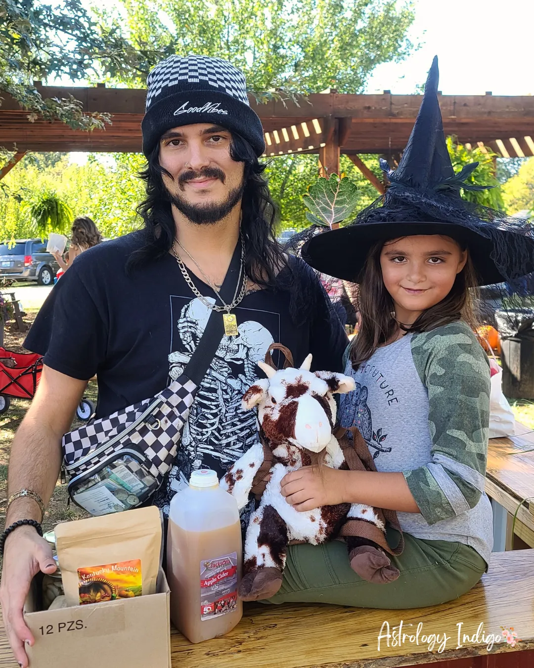 A man stands in a skeleton shirt with his daughter who is wearing a witch hat. They are outside at a pumpkin place.